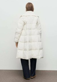 Persona de pie de espaldas, vistiendo un largo abrigo acolchado blanco, pantalones anchos oscuros y zapatos marrones, contra un fondo plano.