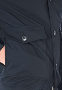 Giacca blu navy realizzata in un materiale sintetico liscio, con chiusura a bottone e una grande tasca a soffietto sul petto per riporre gli oggetti.