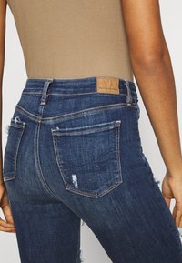 Mörkblå högmidjade jeans med sliten finish, med en tan läderlogotyp, bakfickor och subtila sömnadsdetaljer.