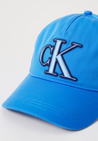 Blauwe pet met een gestructureerd ontwerp, voorzien van een geborduurd "ck" logo in marineblauw. Gemaakt van een gladde stof met een gebogen rand.