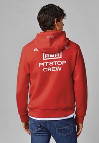 Rdeče kapuco s prilagodljivo vrvico, ki na hrbtni strani nosi bel napis "RBR PIT STOP CREW". Gladka tekstura, sproščen kroj, v kombinaciji z modrimi kavbojkami.
