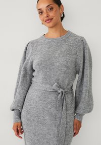 Grijs gebreid kleed met lange pofmouwen en een strikceintuur in de taille. Het heeft een ribbelstructuur over het hele kleed.