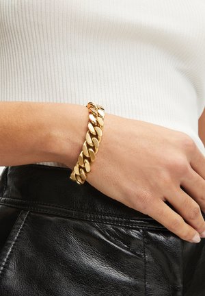 Bracelet en chaîne dorée avec des maillons ovales, finition polie et fermoir. Porté sur un poignet contre un fond en cuir noir.