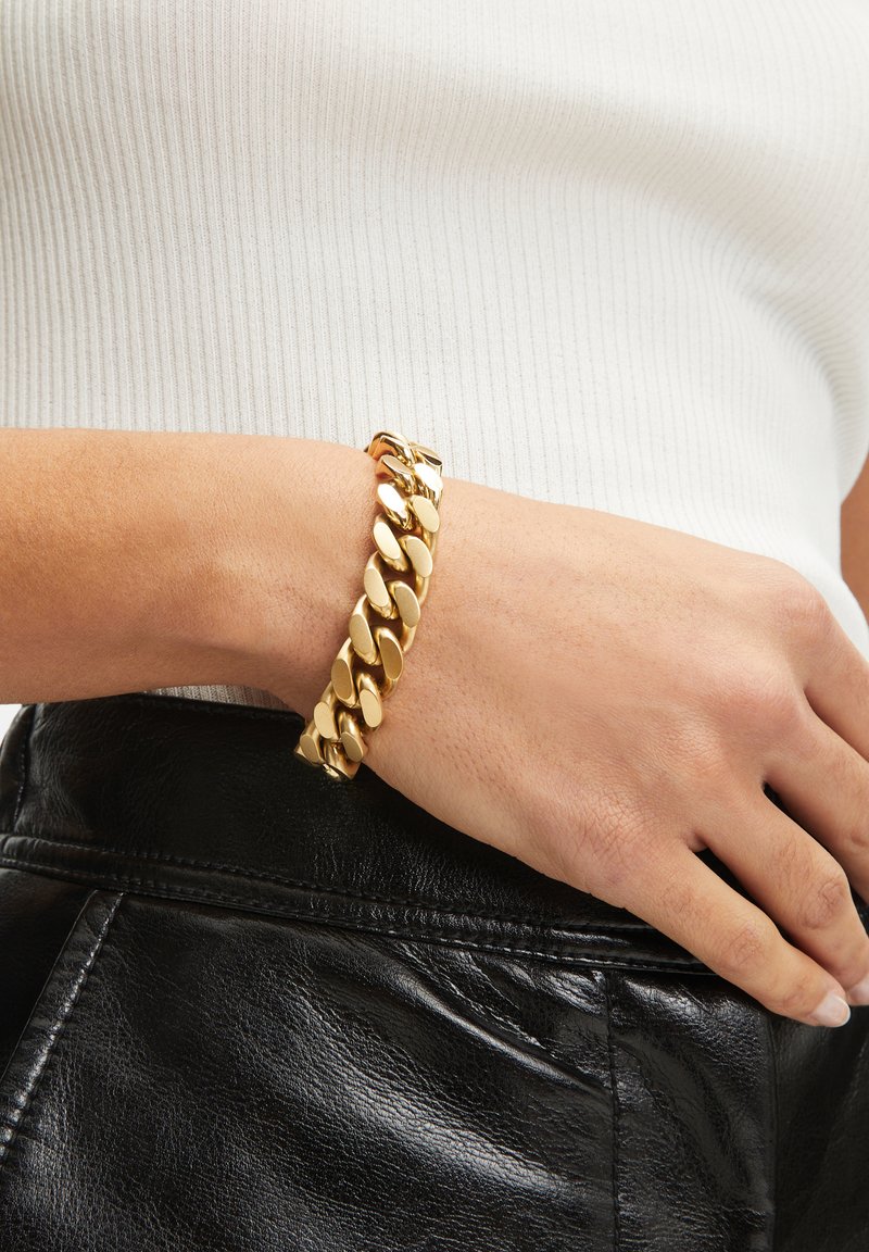 Bracelet en chaîne dorée avec des maillons ovales, finition polie et fermoir. Porté sur un poignet contre un fond en cuir noir.