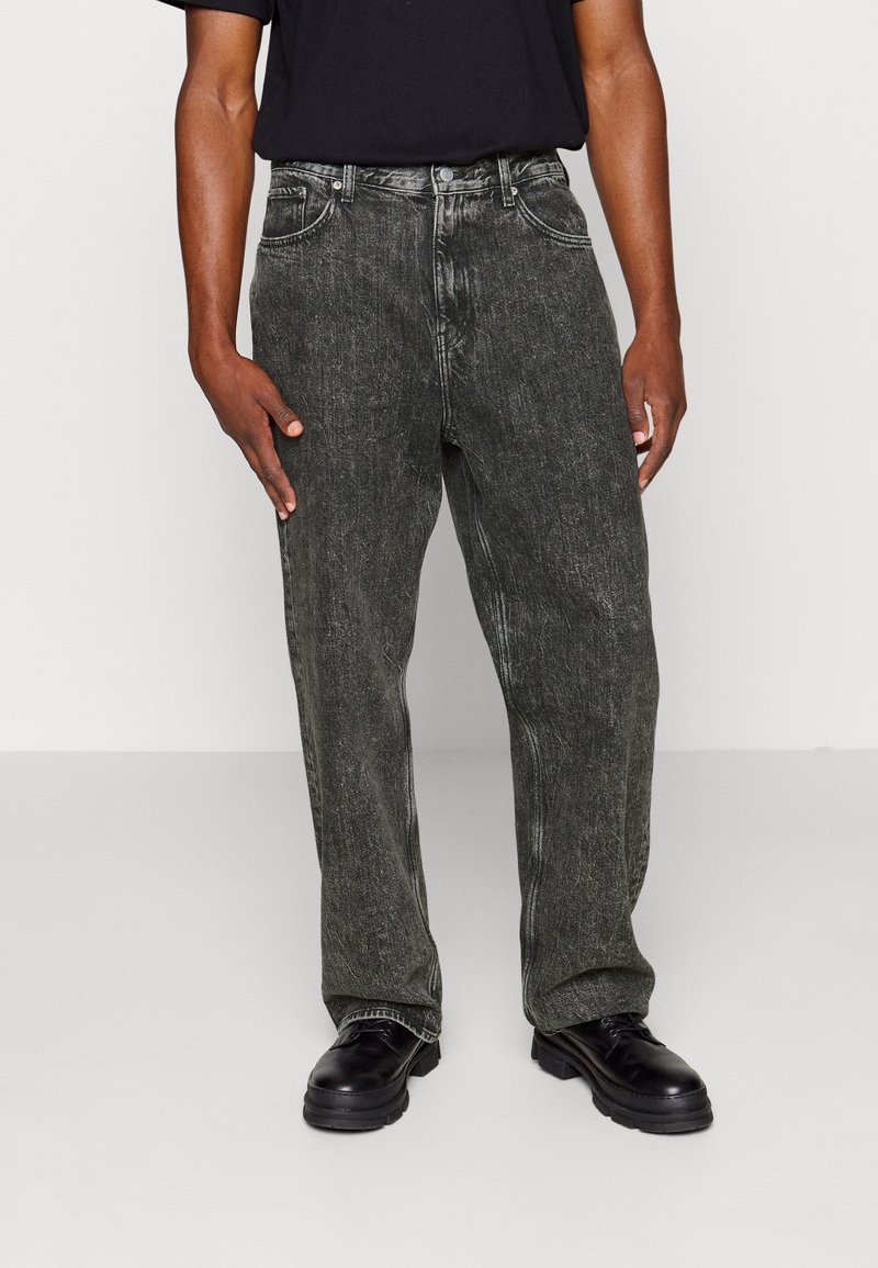 Loose-fit, gray denim jeans with a faded finish, featuring five pockets and contrasting stitching, paired with black shoes.