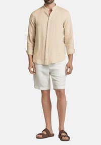 Camicia di lino beige con design senza colletto, pantaloni corti bianchi e sandali marroni. La camicia ha maniche lunghe arrotolate fino al gomito.