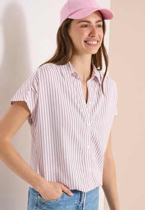 Femme souriante portant une casquette de baseball rose, un chemisier blanc à manches courtes avec de fines rayures rouges et un jean bleu, debout avec une main dans la poche.