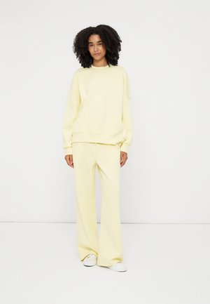 Kvinde med krøllet hår iført en lys gul oversized sweatshirt og matchende vidde bukser med hvide sneakers, står mod en hvid baggrund.