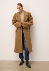 Długi, oversize brązowy płaszcz wełniany z szerokimi klapami, bocznymi kieszeniami i lekko teksturowanym wykończeniem, w połączeniu z niebieskimi dżinsami i czarnymi botkami za kostkę.