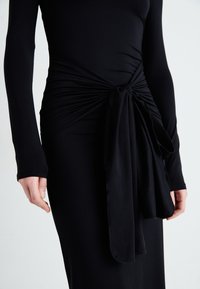 Μαύρο φόρεμα με μακριά μανίκια που διαθέτει λεπτομέρεια με σπαστό κόμπο στη μέση. Μαλακό, υφασμένο ύφασμα με λεία, εφαρμοστή σιλουέτα.
