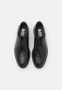 Un paio di scarpe formali in pelle nera lucida con lacci, dotate di solette firmate, esposte affiancate su uno sfondo bianco.