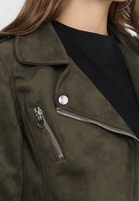 Primo piano di una giacca in suede verde oliva con dettagli in zip e bottoni argentati indossata su una maglietta nera a girocollo.
