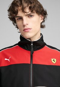 Giacca nera e rossa con zip e colletto alto, con il logo Puma a sinistra e lo stemma Ferrari a destra, in tessuto testurizzato.