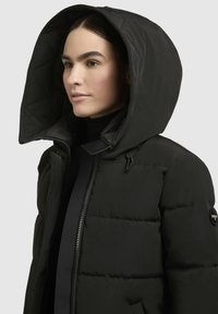 Sieviete, kas valkā melnu pūkotu jakas ar lielu kapuci, daļēji aizvērtu, ar mazu apaļu logotipa uzšuvumu uz piedurknes.