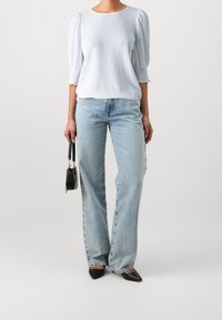 Top azzurro chiaro con texture e maniche a sbuffo 3/4, abbinato a jeans a gamba larga azzurri chiari e ballerine nere a punta. Piccola borsa nera in mano.