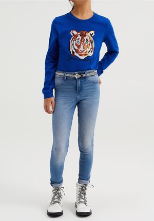 Kind trägt ein blaues Langarmshirt mit Tigerkopf-Design, hellblaue Jeans mit silbernem Gürtel und weiße Schnürstiefel.