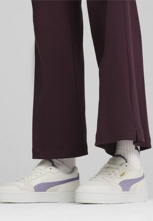 Witte Puma-sneakers met paarse strepen, gedragen met lichtgrijze sokken en donkerpaarse geribbelde wijde pijpen, tegen een lichtgrijze achtergrond.