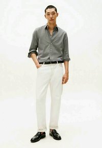 Chemise à rayures grises et blanches, pantalon blanc, ceinture noire et mocassins noirs. Le modèle se tient avec les mains dans les poches devant un fond uni.