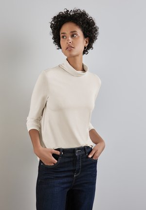 Un haut col montant beige en tissu doux avec des manches longues, associé à un jean en denim noir. Le haut présente une coupe décontractée et un design minimaliste.