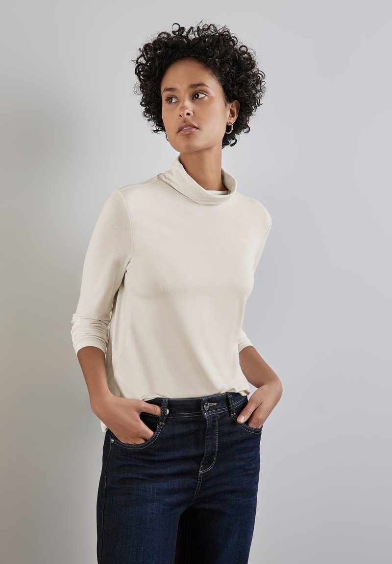 Un haut col montant beige en tissu doux avec des manches longues, associé à un jean en denim noir. Le haut présente une coupe décontractée et un design minimaliste.