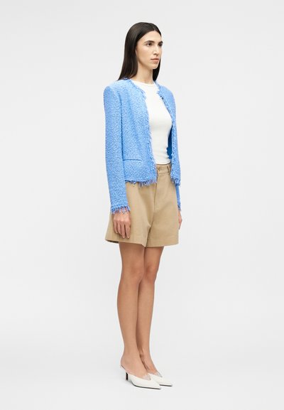 Γυναίκα που φοράει μια υφασμάτινη γαλάζια ζακέτα με κρόσσια, λευκό τοπ, μπεζ σορτς και λευκά μυτερά τακούνια, στέκεται πλάγια σε απλό φόντο.