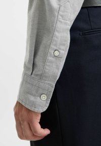 Camicia grigia chiaro con tessuto texturizzato; presenta un polsino a risvolto con bottoni bianchi; indossata con pantaloni scuri.