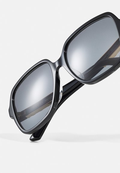 Lunettes de soleil noires surdimensionnées avec des verres gris. La monture présente une finition lisse et brillante avec un design angulaire distinctif.