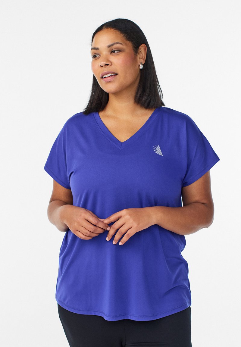 Femme aux cheveux bruns mi-longs portant un t-shirt bleu à encolure en V et des boucles d'oreilles en argent, debout les mains légèrement jointes.