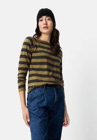 Gestreiftes Langarmshirt in Olive und Navy, kombiniert mit hoch taillierten blauen Jeanshosen. Das Model trägt eine schwarze Mütze und präsentiert eine legere Passform.