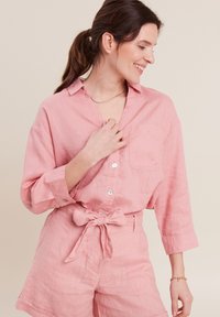 Camicia di lino rosa chiaro con colletto, chiusura con bottoni e taschino sul petto, abbinata a pantaloni corti a vita alta coordinati e una cintura annodata.