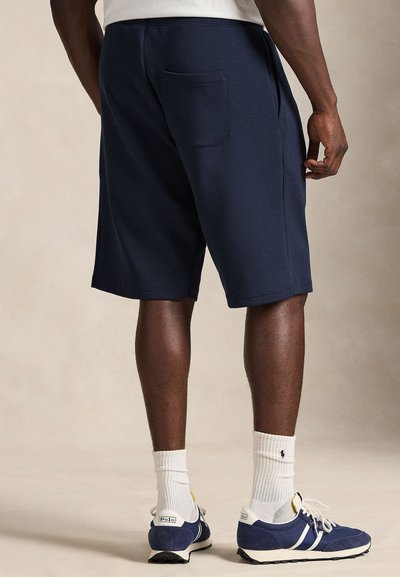 Οι σκούρο μπλε βερμούδες από φούτερ διαθέτουν πλαϊνές και πίσω τσέπες. Φοριούνται με λευκές κάλτσες και μπλε αθλητικά παπούτσια με λευκές λεπτομέρειες και λογότυπο.