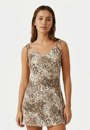 Žena oblečená v bezrukávových béžových minišatách s čiernym a hnedým zvieracím vzorom a zaväzovacími ramienkami na jednoduchom bielom pozadí.