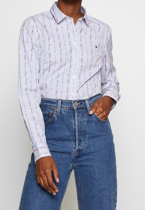 Personne portant une chemise blanche à boutons avec des rayures verticales bleues et rouges, rentrée dans un jean bleu taille haute, une main dans la poche.