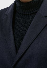 Blazer azul marino en tejido suave con solapas de muesca, superpuesto sobre un suéter de cuello alto acanalado. El tejido texturizado aporta dimensión y contraste.