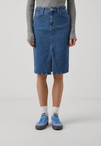 Jupe en denim bleu moyen, longueur genou avec une fente avant, taille haute et design à cinq poches. Portée avec des baskets bleu clair et des chaussettes blanches.