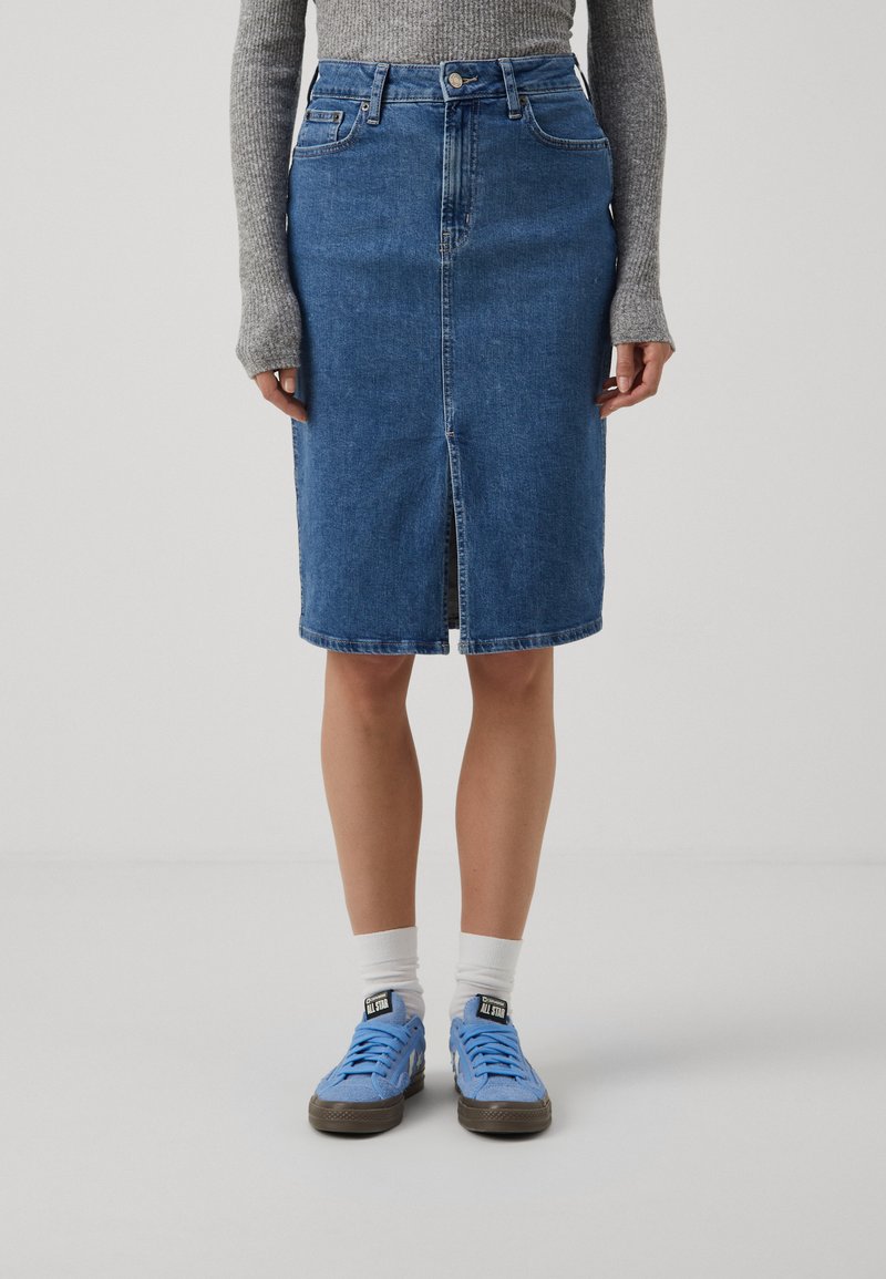 Jupe en denim bleu moyen, longueur genou avec une fente avant, taille haute et design à cinq poches. Portée avec des baskets bleu clair et des chaussettes blanches.