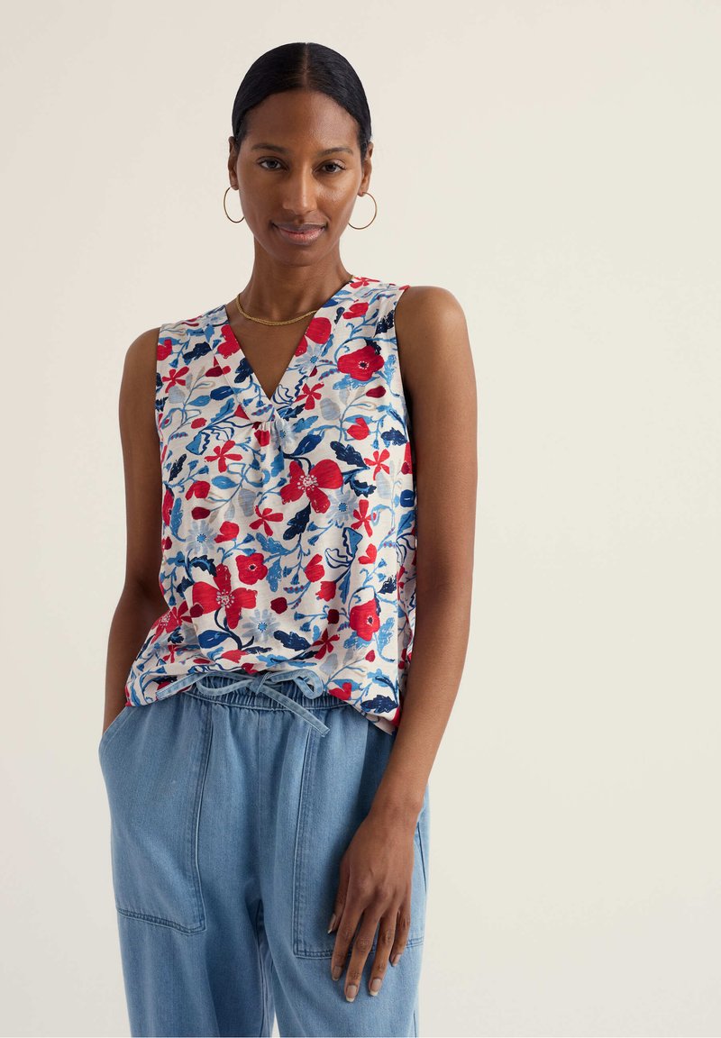 Mujer con cabello oscuro recogido, lleva pendientes de aro dorados, blusa sin mangas con estampado floral en rojo, azul y blanco, y pantalones de tiro alto azul claro con cordón, con las manos en los bolsillos.