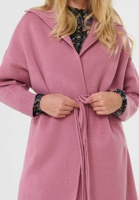 Femme attachant la ceinture à la taille d'un long cardigan en maille rose sur un haut à fleurs sombre, avec des cheveux blonds visibles.