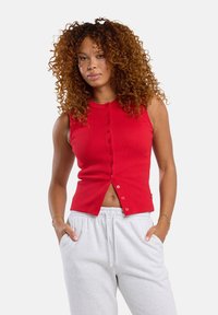 Red sleeveless top made of ribbed fabric, featuring a buttoned front and a slightly cropped hem. Worn with gray sweatpants.