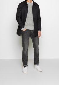 Muž v černém kabátu, šedém svetru, tmavě šedých džínách a bílých teniskách stojící s jednou rukou v kapse na bílém pozadí.