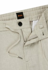 Beige linen trousers featuring a black button, zip fly, drawstring, and a label with "BOSS" in orange. Smooth texture and simple design.