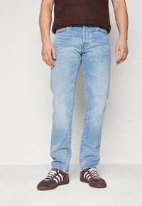 Vaqueros de mezclilla azul claro con corte recto, que presentan un diseño clásico de cinco bolsillos y un ligero desgaste. El modelo lleva zapatillas oscuras con rayas.