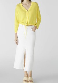 Gelbe, leichte Strickjacke mit V-Ausschnitt und Knöpfen, kombiniert mit einem weißen, knöchellangen Jeansrock mit einem Schlitz vorne.