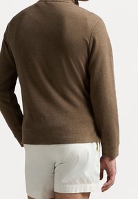 Braunes Langarmoberteil aus strukturiertem Stoff mit rundem Halsausschnitt und engen Bündchen, kombiniert mit hellen Shorts.