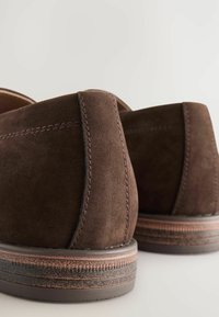 Zapatos de ante marrón, con una textura suave, parte trasera redondeada y detalles de costura en contraste, con suela de goma y un sutil patrón de tracción.