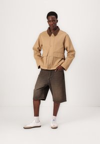 Beige canvas jacket with brown collar, two front pockets, and button closure, paired with dark denim shorts and white sneakers.