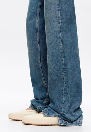 Scarpe slip-on beige chiaro con suole testurizzate, abbinate a jeans in denim blu a vestibilità rilassata, con dettagli a orlo sfilacciato, mostrate dalla caviglia in giù.