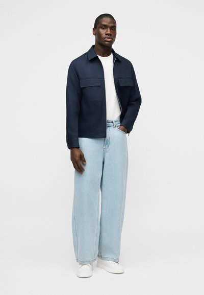 Νεαρός άνδρας όρθιος, φοράει σκούρο μπλε σακάκι, λευκό μπλουζάκι, ανοιχτό μπλε τζιν με φαρδιά πόδια και λευκά αθλητικά παπούτσια με φόντο απλό.