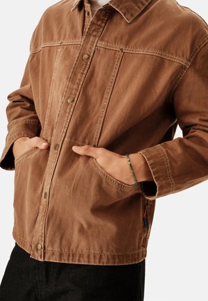 Chaqueta de mezclilla marrón con cuello, cierres frontales de broches, bolsillos laterales y detalles de costuras visibles. Presenta un diseño ligeramente recortado.