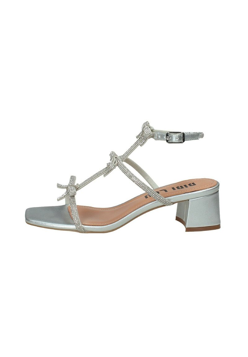 Sandalo argentato con cinturini e punta quadrata, con tacco basso a blocco. Caratterizzato da fiocchi decorativi e chiusura con fibbia. Materiale sintetico liscio.